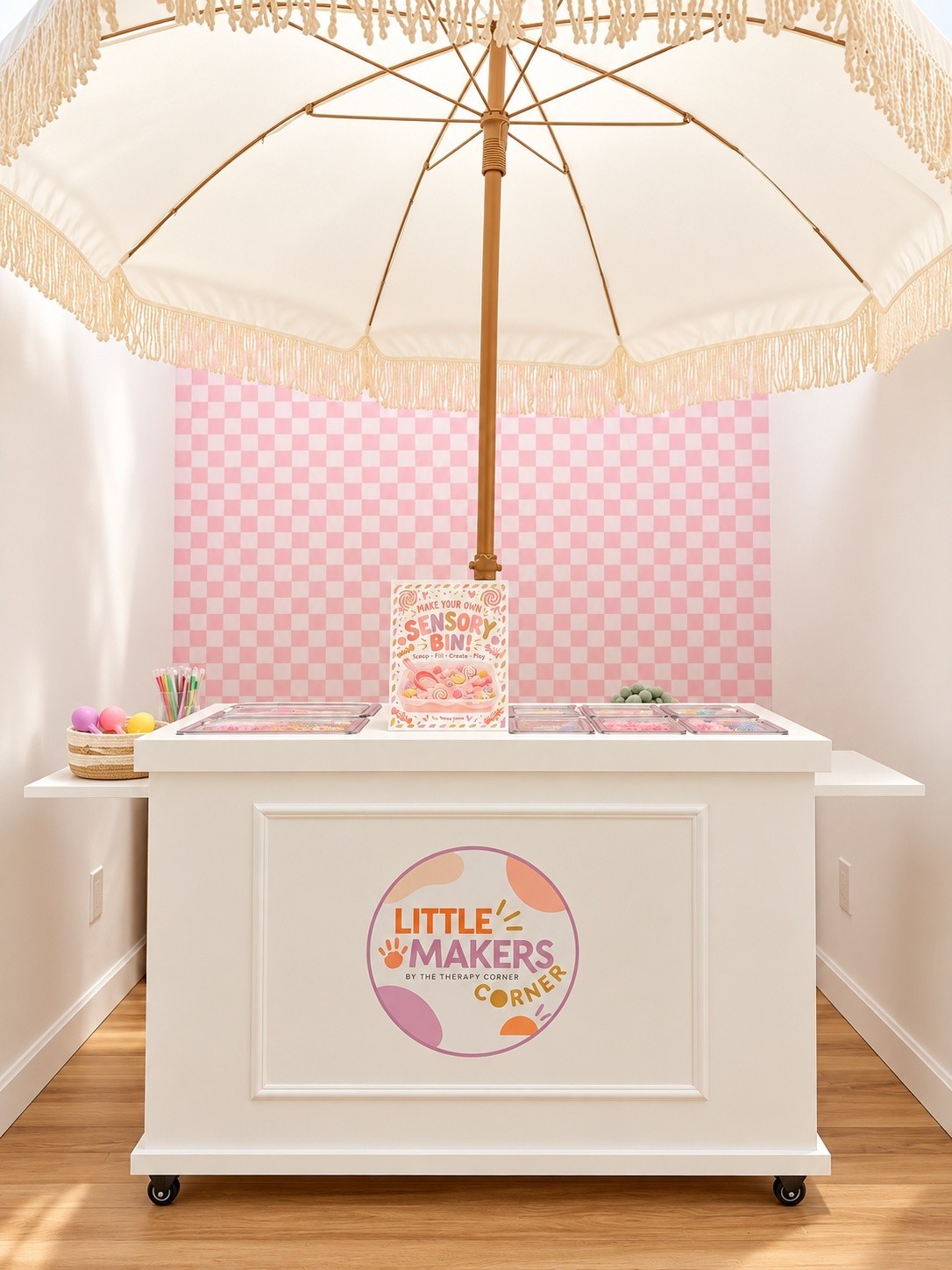 Little Makers Corner activity cart with a fringed umbrella, pink checkerboard backdrop, and sensory-bin supplies