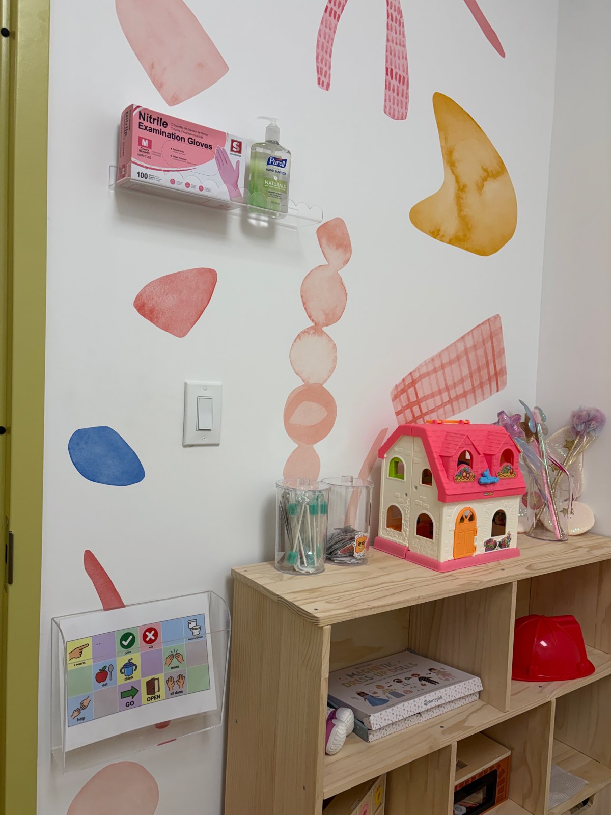 Colorful abstract wall shapes above wooden toy shelves and a dollhouse in the clinic